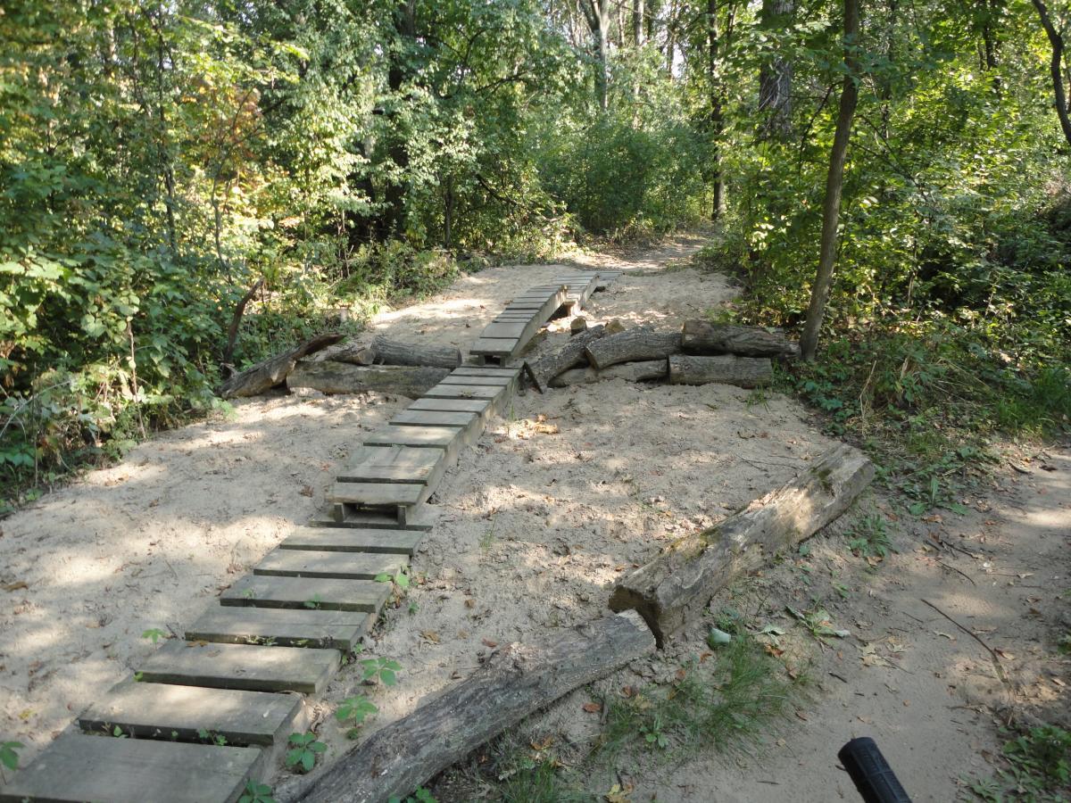 A narrow wooden bridge crossing a sandy path in a wooded area, surrounded by greenery and trees. Hillside Park mountain bike trail.