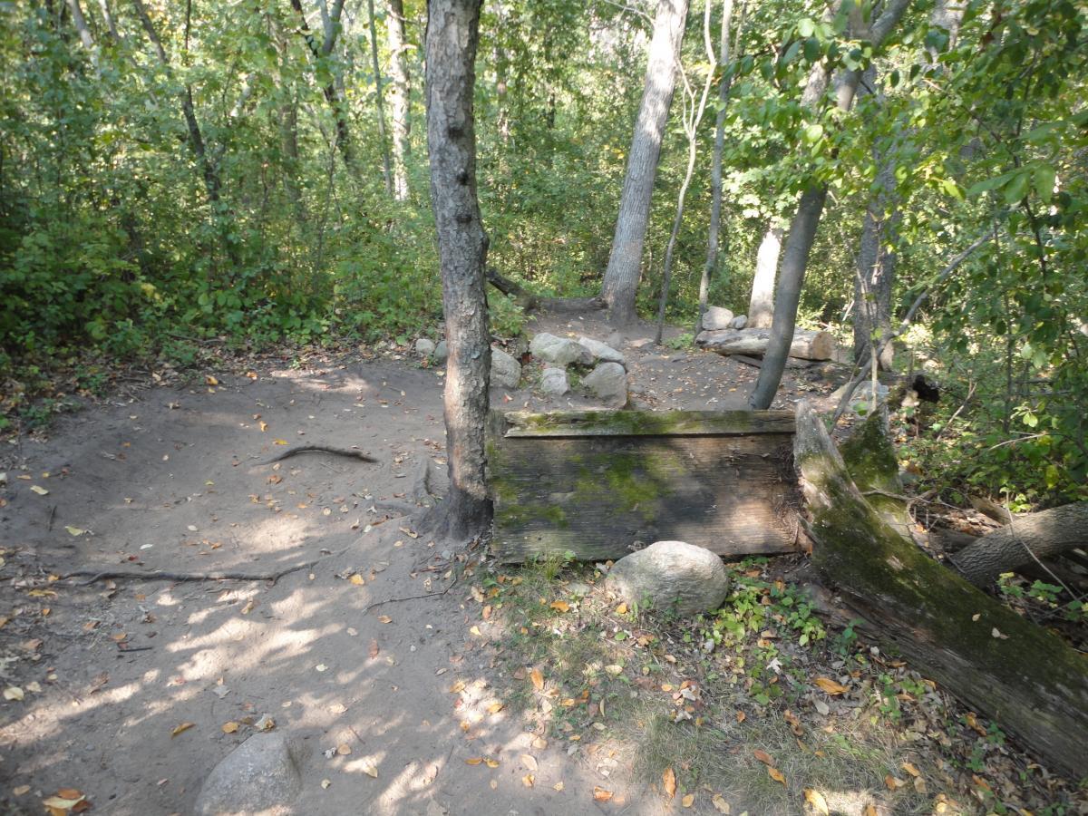 A narrow path in a wooded area, surrounded by green trees and scattered rocks. The ground is sandy with some exposed roots and fallen leaves, indicating a natural, earthy setting. A wooden structure is partially visible, blending into the landscape. Hillside Park mountain bike trail.