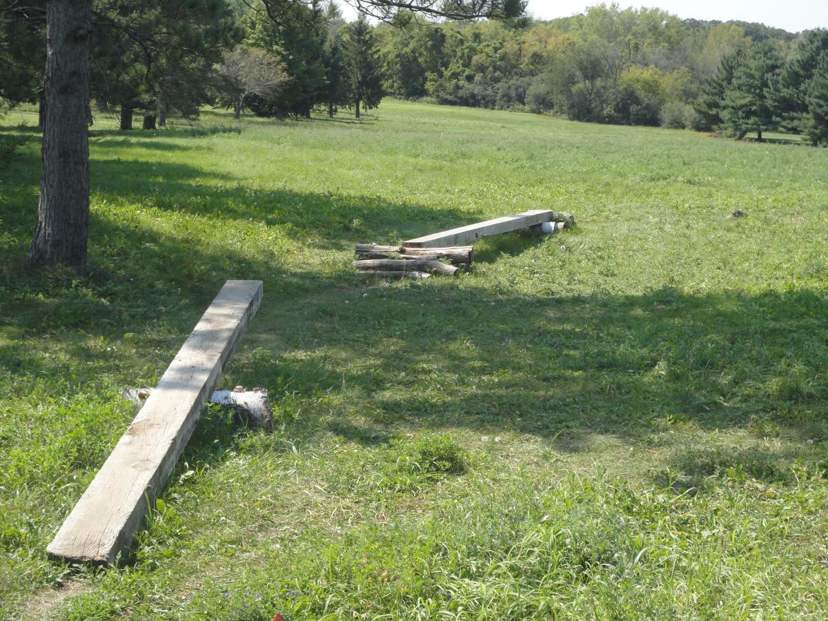 A grassy field with wooden planks lying on the ground, some positioned horizontally and others stacked. Surrounding trees provide a natural backdrop to the scene. Eastwood Trails mountain bike trail.