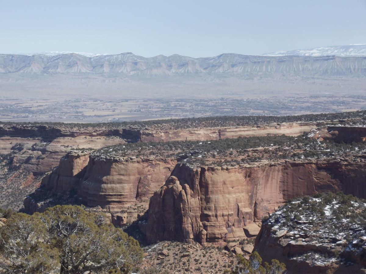 A panoramic view of layered red rock formations and distant mountains under a clear blue sky, showcasing the natural beauty of a canyon landscape. The foreground features rugged cliffs and patches of snow, while the rolling hills and mountains in the background create a stunning backdrop. Colorado National Monument mountain bike trail.