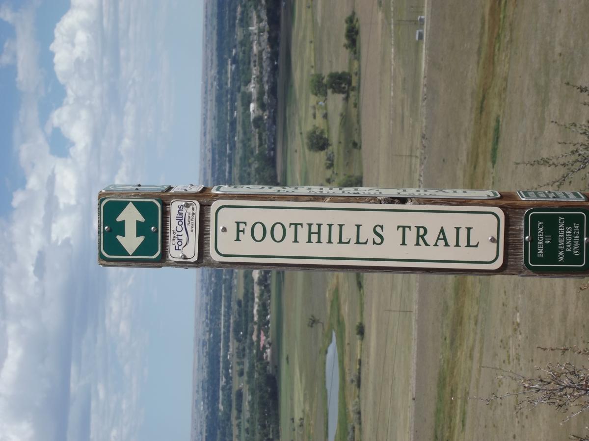 Signpost indicating the Foothills Trail, featuring directional arrows and additional information about emergency contacts. The background includes a scenic view of fields and distant hills under a partly cloudy sky. Foothills Trail mountain bike trail.