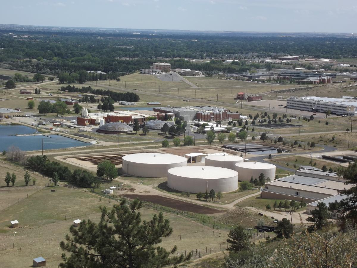 Aerial view of a large industrial complex featuring several circular storage tanks, various buildings, and a small pond. The surrounding landscape includes grassland and trees, with a distant view of more buildings and rolling hills under a partly cloudy sky. Foothills Trail mountain bike trail.