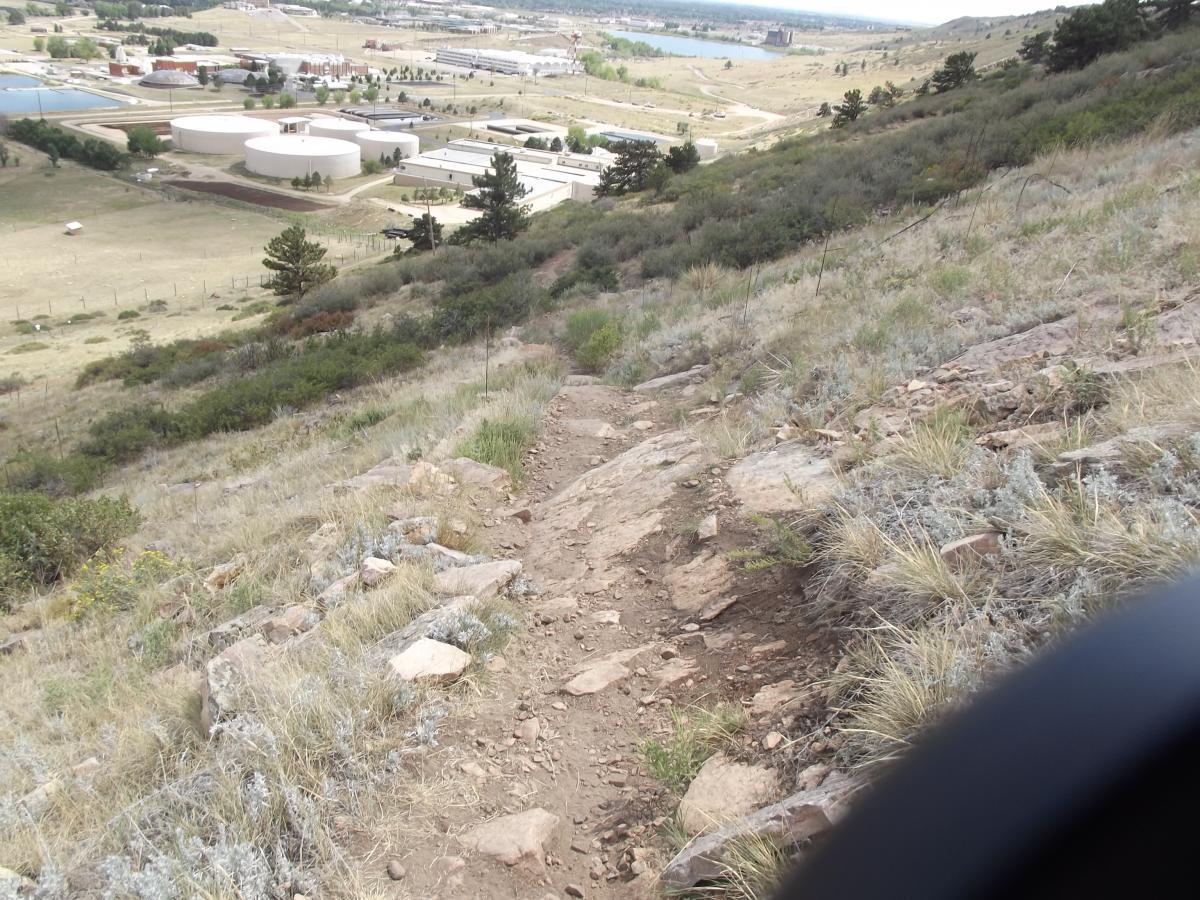 A rocky hiking trail descending a grassy hillside, with a view of a valley below that features several industrial buildings, water tanks, and a large body of water in the distance under a cloudy sky. Foothills Trail mountain bike trail.