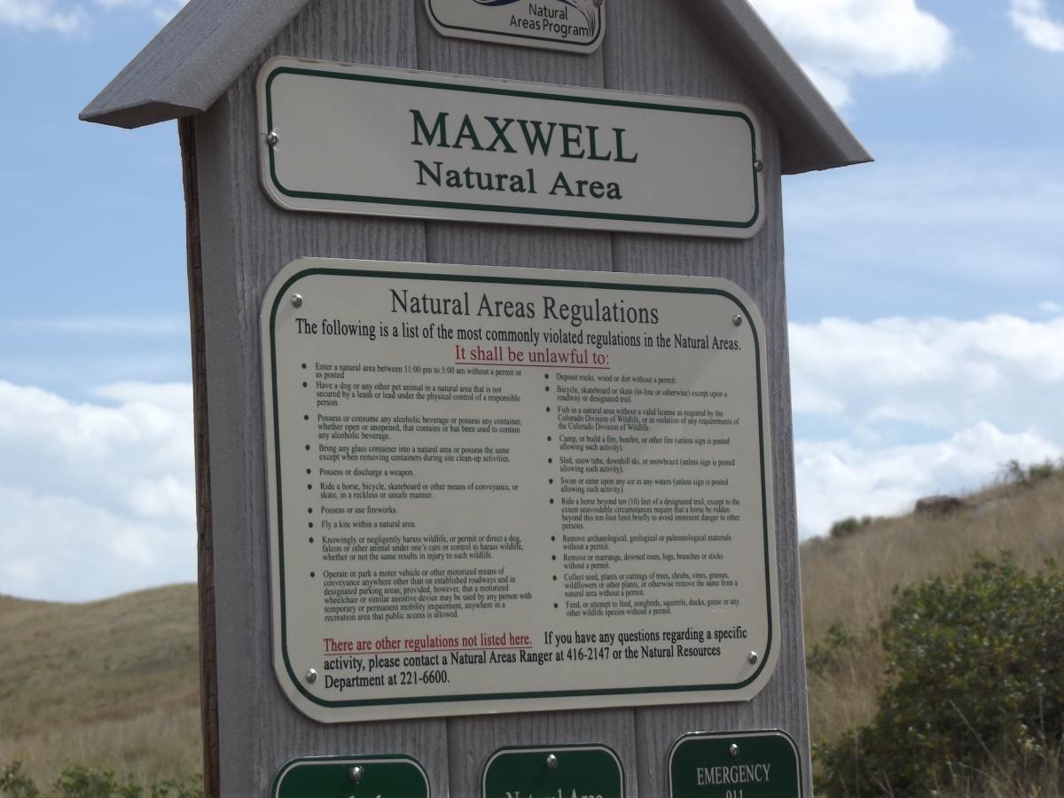 Sign for Maxwell Natural Area displaying regulations and guidelines for visitors. The sign lists activities that are prohibited in the area, such as entering without a permit, consuming alcohol, and using weapons. It also provides contact information for additional inquiries. The background features natural scenery with a few clouds in the sky. Maxwell's mountain bike trail.