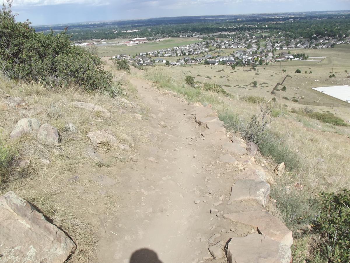 A rocky hiking trail descends from a hillside, with grass and shrubs bordering the path. Below, a scenic view reveals a sprawling landscape of homes, fields, and distant hills under a partly cloudy sky. Maxwell's mountain bike trail.