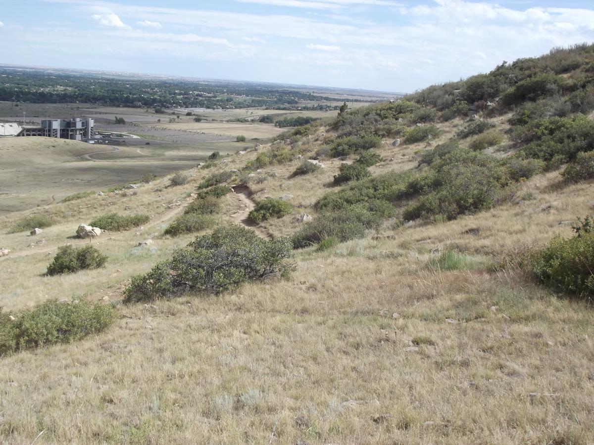 A scenic view of a grassy hillside with sparse shrubs and rocks, leading down to a flat landscape. In the background, a modern building is visible along with a paved area. The sky is partly cloudy, and the horizon stretches out over open fields and distant trees. Maxwell's mountain bike trail.