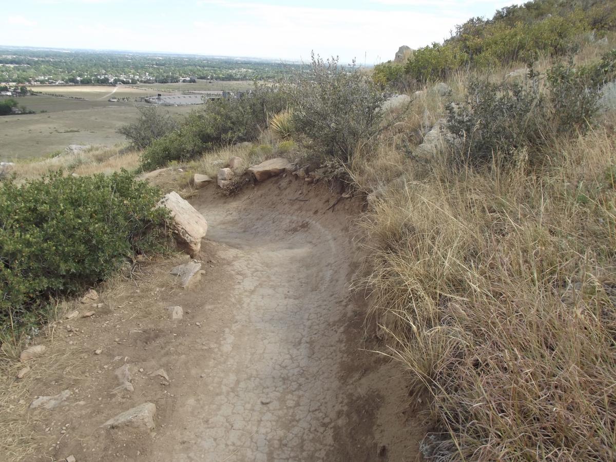 A winding dirt path traverses a hillside, surrounded by patches of dry grass and shrubs. In the background, a vista of distant fields and a small town is visible under a partly cloudy sky. Natural stone formations line the side of the trail, indicating a rugged outdoor setting ideal for hiking or biking. Maxwell's mountain bike trail.