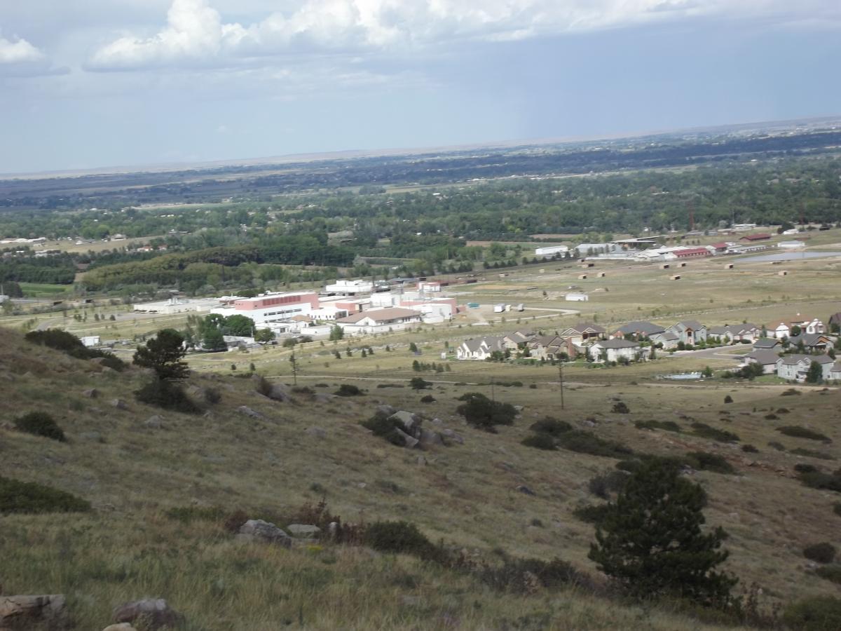 A panoramic view of a rural landscape featuring a mix of open fields, residential homes, and commercial buildings. The foreground shows grassy hills with boulders, while the background reveals a small town situated among trees and fields, under a cloudy sky. Maxwell's mountain bike trail.