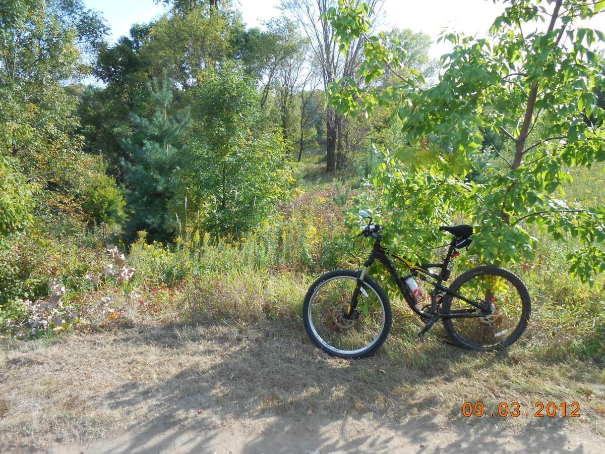 A black mountain bike is parked beside a grassy area, surrounded by various trees and green foliage. The scene is illuminated by sunlight, showcasing the vibrant colors of nature. A date stamp in the lower right corner reads "09.03.2012." Salem Hills Mountain Bike Trail mountain bike trail.