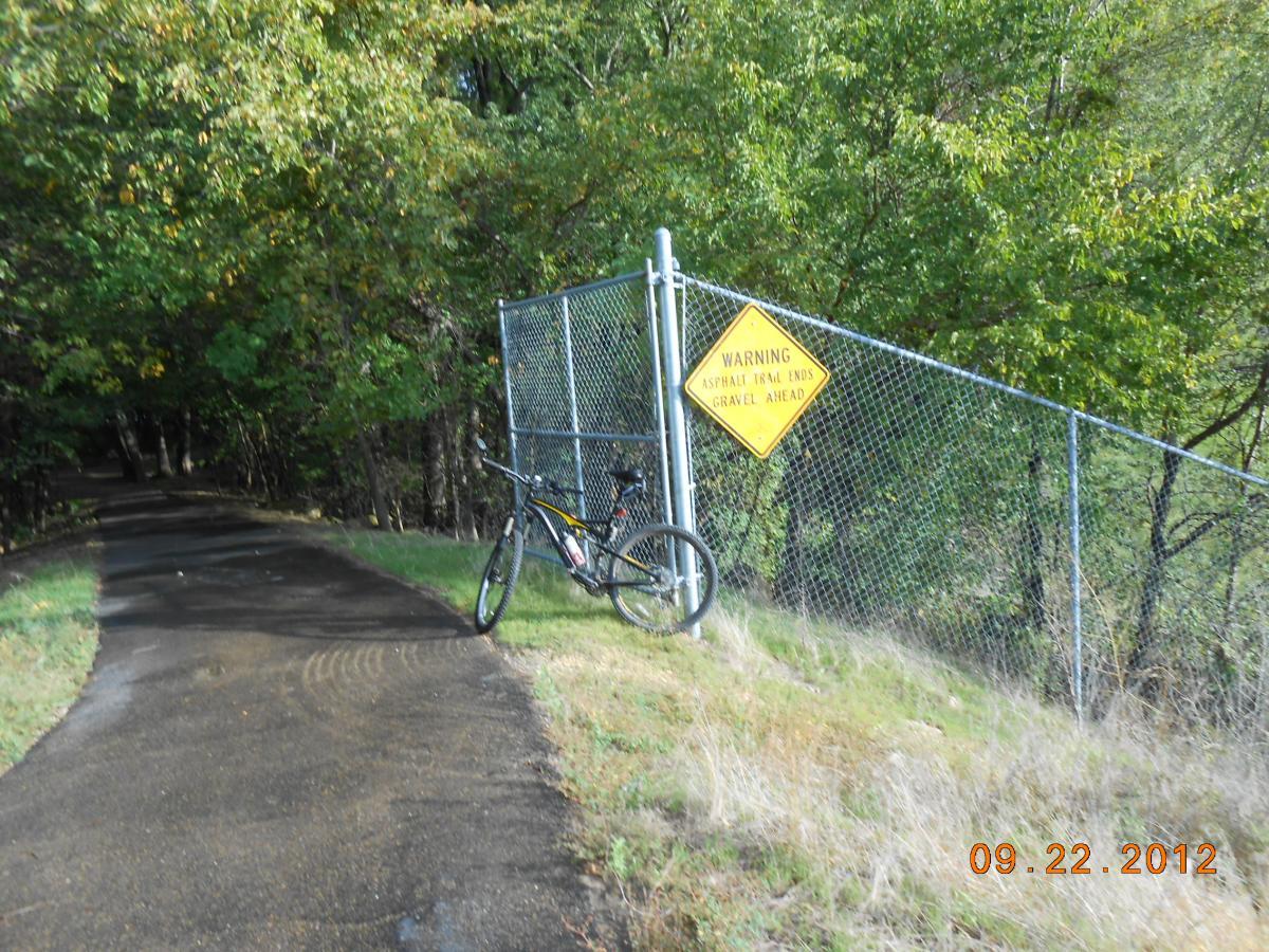 A mountain bike rests against a chain-link fence next to a paved path. A yellow warning sign states, "WARNING: Asphalt Trail Ends, Gravel Ahead," surrounded by trees in a natural setting. The image captures the transition from a smooth trail to a gravel area. Discovery Trail mountain bike trail.