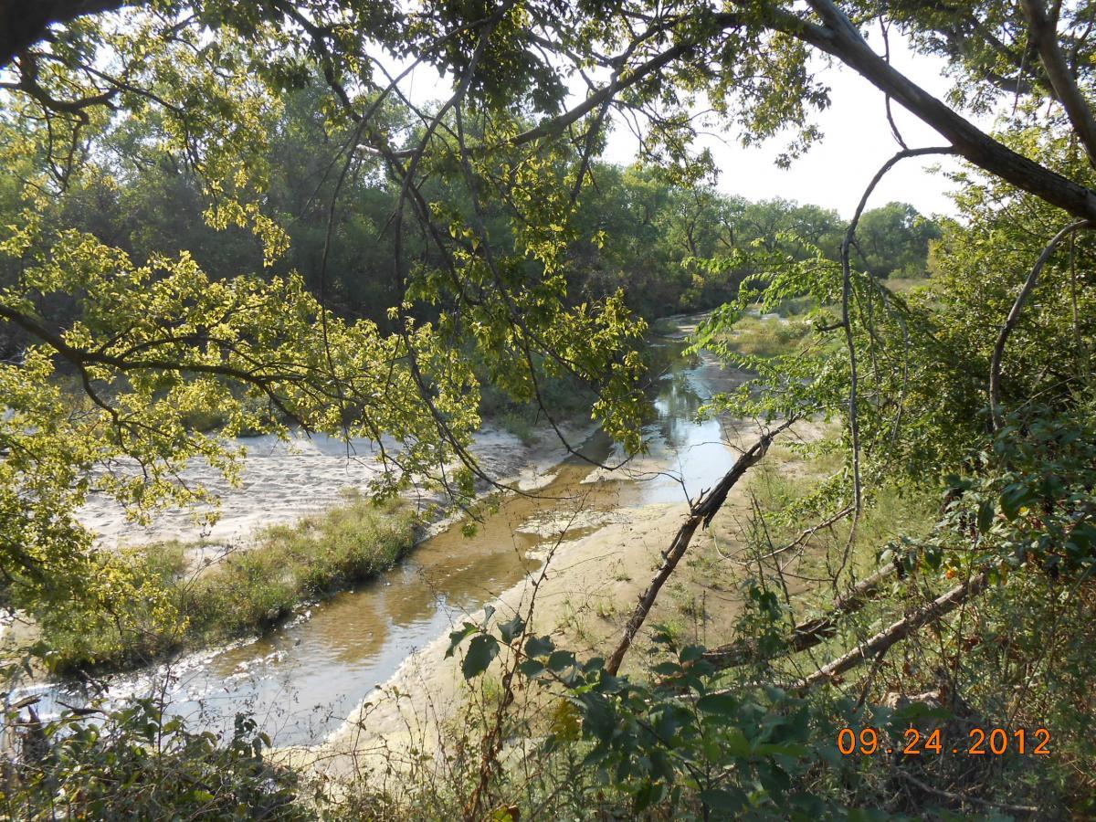 A serene river landscape framed by greenery, featuring gently flowing water and sandy banks. Sunlight filters through the leaves, illuminating the scene in a peaceful, natural setting. The image is dated September 24, 2012. Stinchcomb East mountain bike trail.