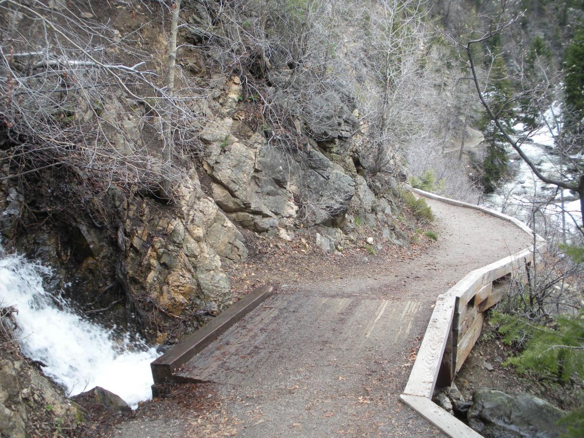 A winding hiking trail alongside a rocky hillside, with a small stream flowing next to it. The path features a wooden railing and is surrounded by sparse trees and bare branches, indicating an early spring or late fall setting. Fish Creek Falls mountain bike trail.