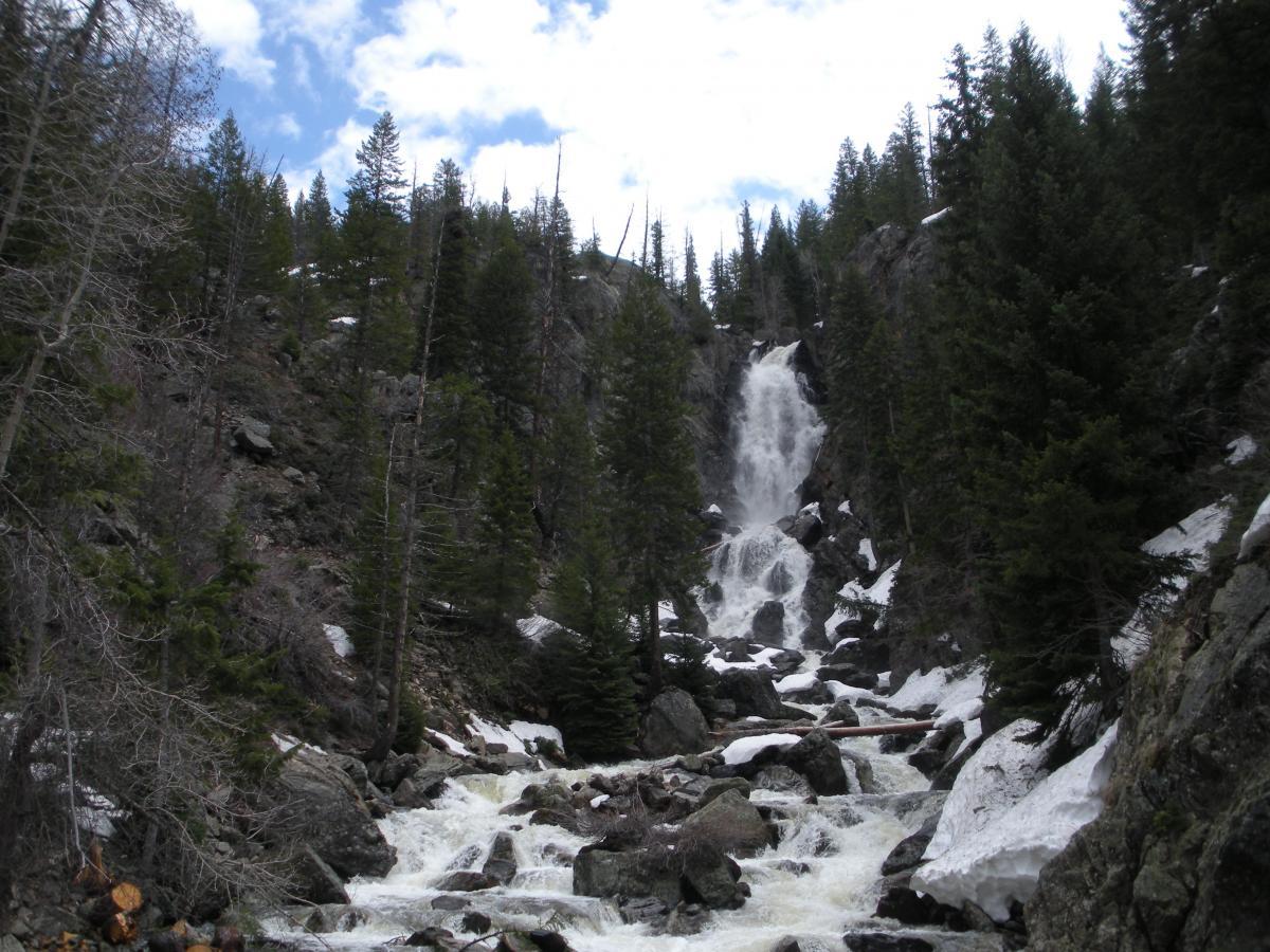 A cascading waterfall flows down a rocky cliff surrounded by evergreen trees, with patches of snow visible on the ground and rocks. The sky is partly cloudy, creating a serene natural landscape. Fish Creek Falls mountain bike trail.