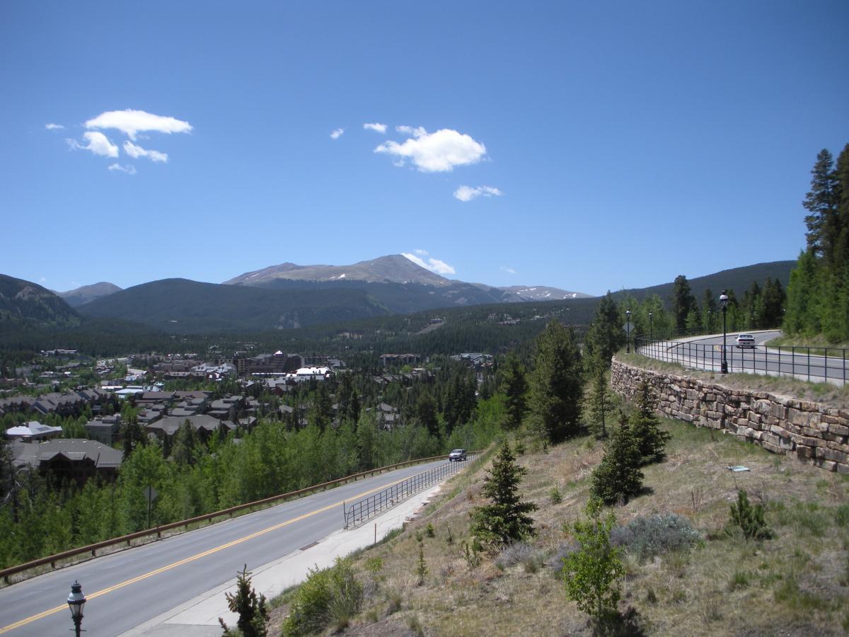 A panoramic view of a mountainous landscape under a clear blue sky, featuring lush green trees and a town nestled in the valley below. A winding road leads up the hillside, alongside a stone wall, with a few scattered clouds in the sky and distant mountains in the background. Peaks Trail mountain bike trail.
