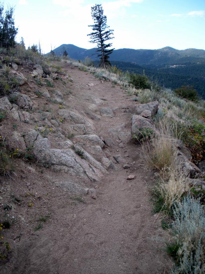 A winding dirt trail lined with rocky outcrops and green grass, leading through a mountainous landscape with distant hills under a partly cloudy sky. Walker Ranch mountain bike trail.