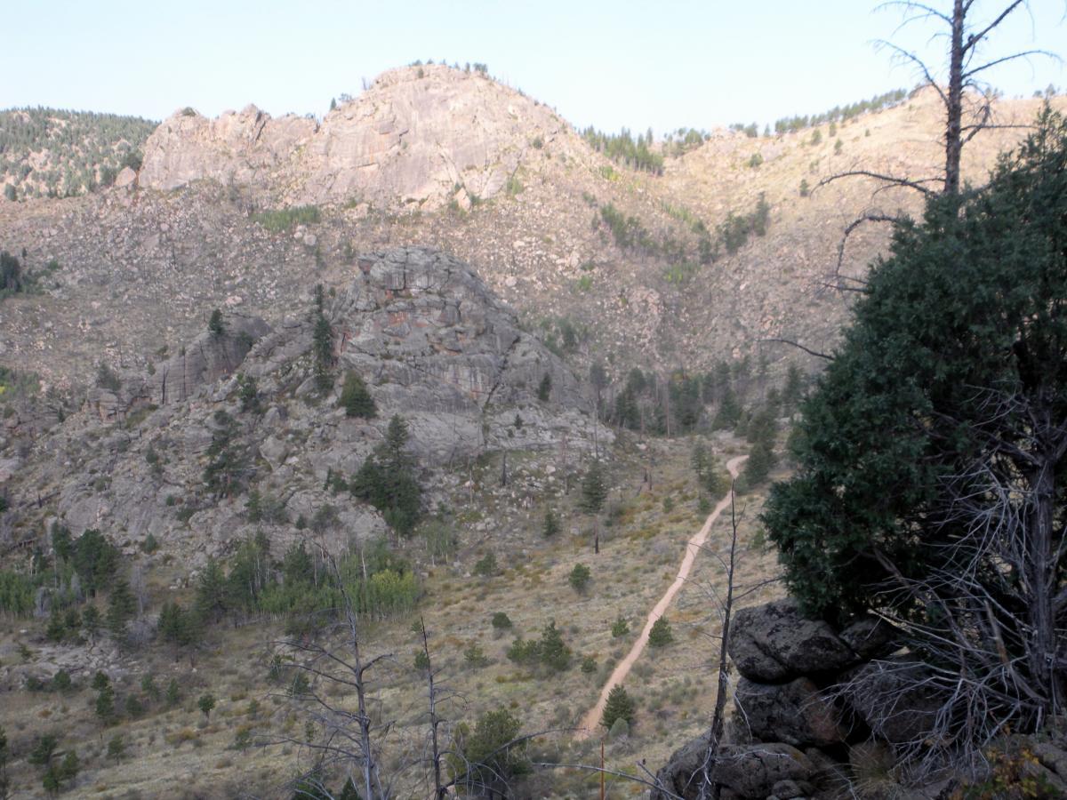 A rocky mountain landscape featuring steep hills, scattered trees, and a winding dirt path that stretches into the distance under a clear sky. The terrain is rugged, with rocky outcrops and varied vegetation throughout the scene. Walker Ranch mountain bike trail.