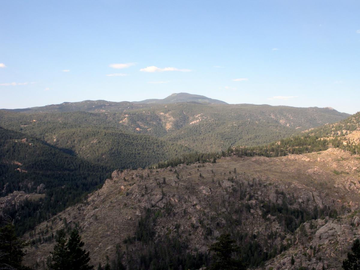 A panoramic view of a mountainous landscape featuring rolling hills covered in dense green forests, rocky outcrops, and a clear blue sky with a few wispy clouds. The foreground shows rugged terrain, while the background includes distant peaks under soft natural lighting. Walker Ranch mountain bike trail.