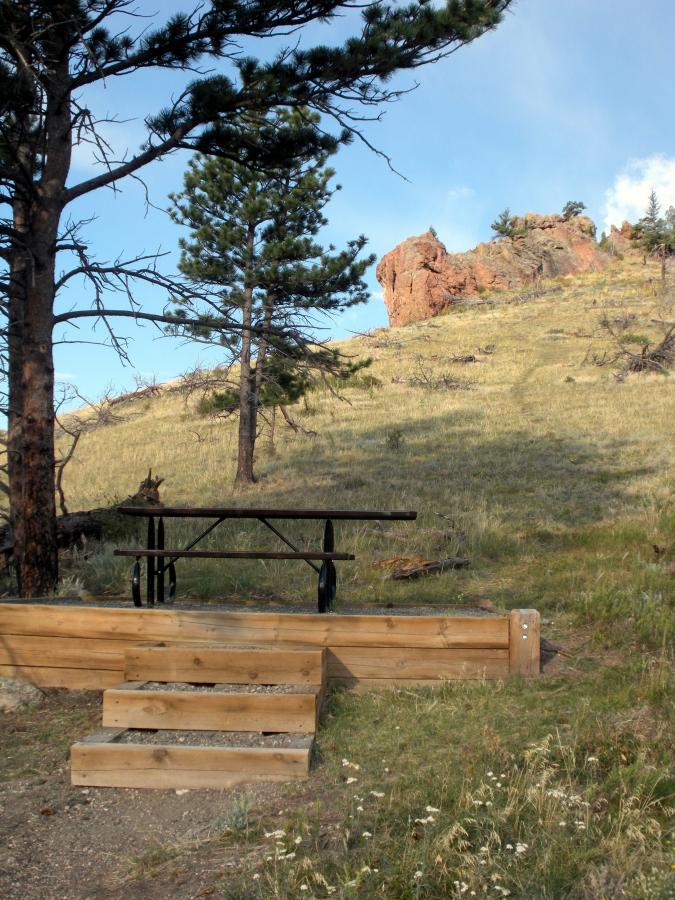 A picnic area featuring a wooden table and stairs, surrounded by trees and a sloping grassy hillside. In the background, rocky outcrops are visible against a blue sky. Walker Ranch mountain bike trail.
