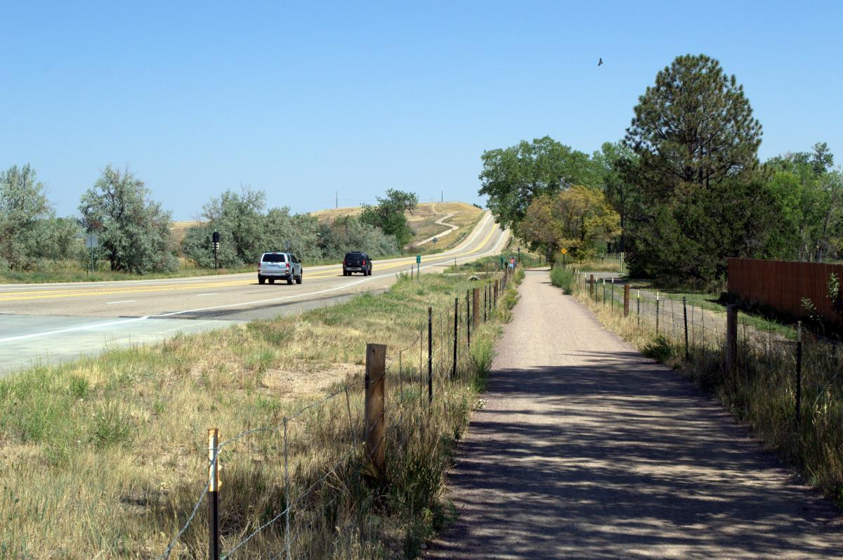 A divided road with two lanes of traffic in each direction, bordered by grassy areas and trees. On the right, a dirt path runs parallel to the road, with a few pedestrians walking nearby. In the distance, the road curves up a hill under a clear blue sky. Coal Creek Trail mountain bike trail.