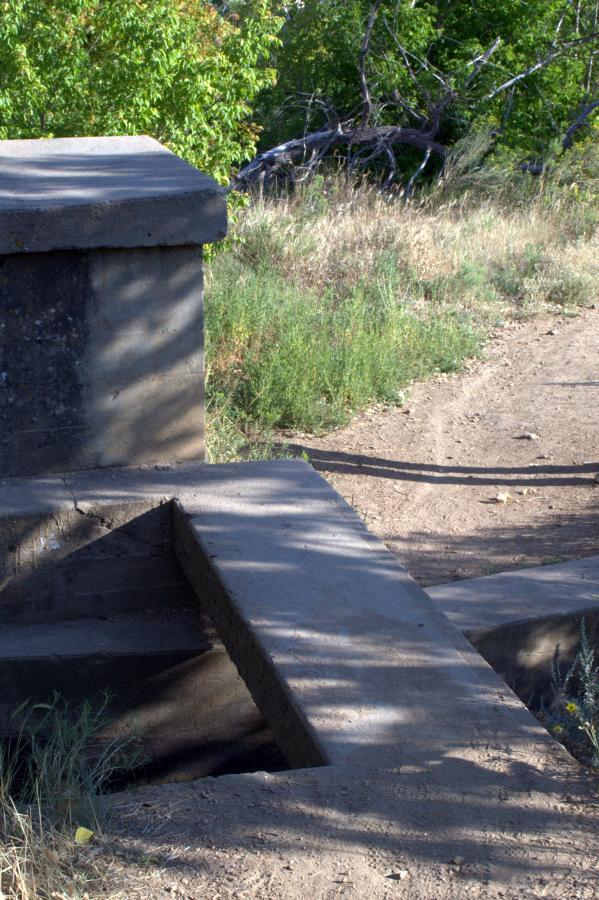 A concrete structure with a sloped surface leading down, surrounded by foliage and a dirt path. The scene is set in a natural environment with green trees and grass in the background. Chimney Gulch mountain bike trail.