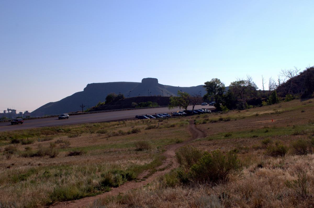 A wide view of a desert landscape featuring a distant mesa under a clear blue sky. In the foreground, a grassy area leads to a dirt path, with several parked cars visible along a road. The mesa is prominent in the background, adding to the natural scenery. Chimney Gulch mountain bike trail.