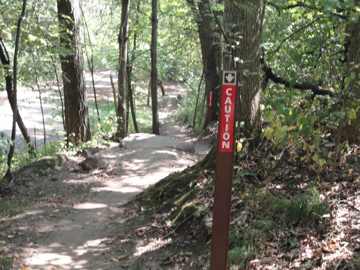 A dirt hiking path surrounded by trees, featuring a prominently displayed caution sign on the right side. The path appears uneven, with rocks and foliage visible, indicating a natural setting. Sunlight filters through the leaves, creating a dappled effect on the ground. Hillside Park mountain bike trail.