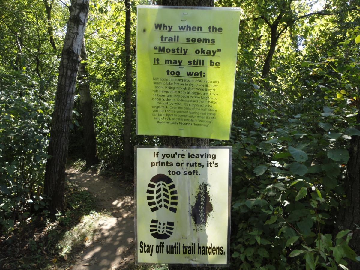 A forest trail sign in a wooded area, featuring two notices. The top sign explains why trails can remain wet even when they appear mostly dry, discussing how soft spots can affect trail conditions. The bottom sign advises trail users to avoid walking on overly soft areas to prevent leaving prints or ruts, stating, "Stay off until trail hardens." The surrounding greenery and natural path are visible, indicating a maintained outdoor trail. Hillside Park mountain bike trail.