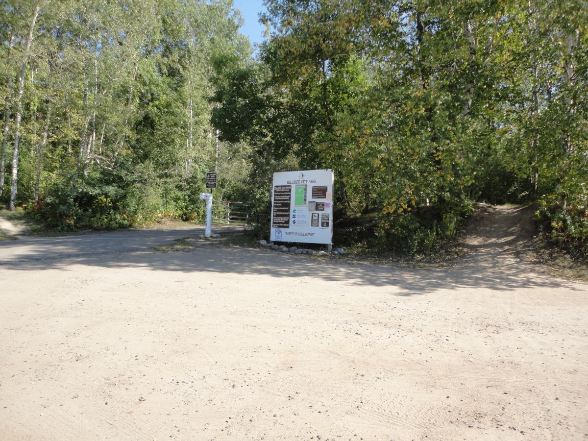 A dirt road intersection leading into a wooded area, with a large informational sign for a city park prominently displayed. The sign provides details about park rules and amenities. Surrounding greenery includes various trees and plants, indicating a natural setting. Hillside Park mountain bike trail.