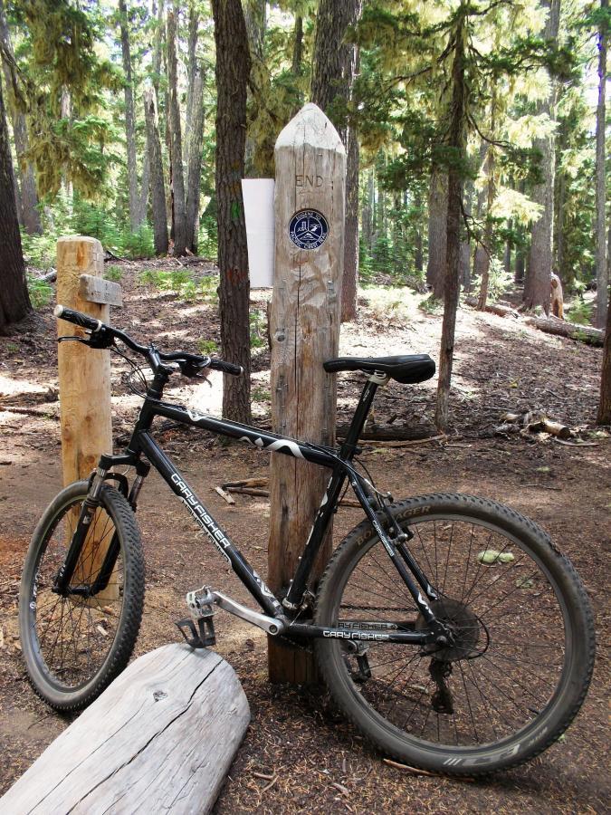 A black mountain bike leaning against a wooden post marked "END" in a wooded area, surrounded by tall trees and forest undergrowth. A small plaque is visible on the post. Gold Lake To Bobby Lake mountain bike trail.