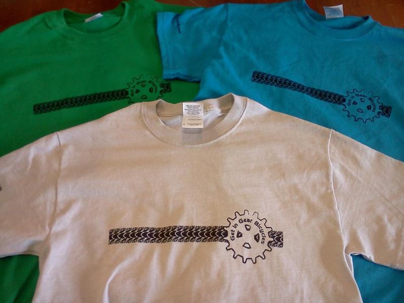 Three t-shirts displayed together. The shirts are in green, blue, and gray. Each shirt features a design with a gear and a tire tread graphic, along with the text "Get in Gear Bicycles." The shirts are laid out on a wooden surface.