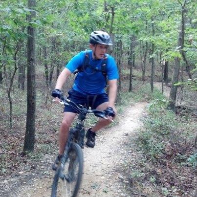 A person wearing a helmet and blue athletic clothing is riding a mountain bike on a dirt trail surrounded by trees. The cyclist is in motion, navigating through a wooded area with greenery in the background. Glacier Ridge Preserve mountain bike trail.