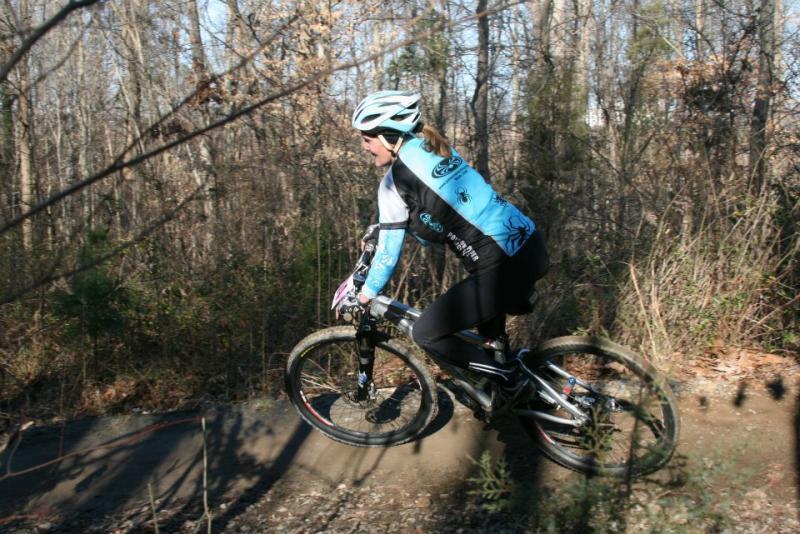 A cyclist riding a mountain bike through a wooded trail, wearing a blue and black cycling jersey and helmet, with trees and foliage in the background.