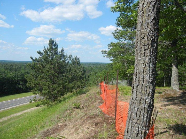 A scenic view from a hillside overlooking a highway, with trees in the foreground and a bright sky with fluffy clouds. An orange safety fence is visible along the edge of the slope, indicating possible construction or maintenance. Hanson Hills mountain bike trail.