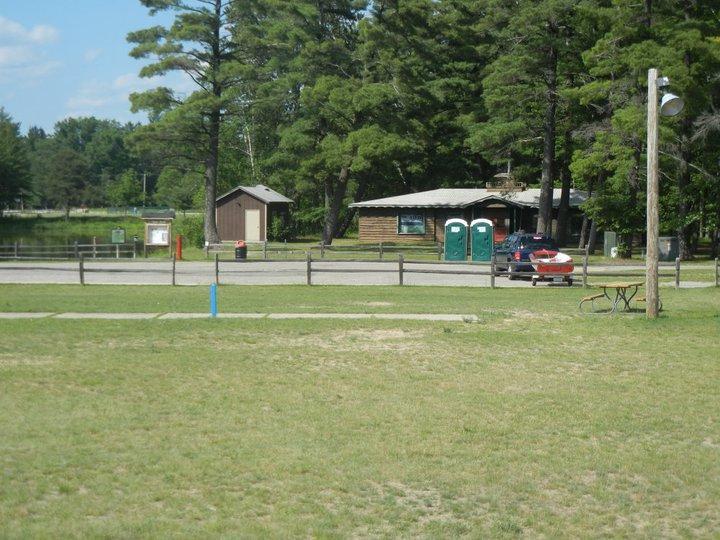 A grassy area in front of a rustic building surrounded by trees, with two portable restrooms visible nearby. In the parking area, a car is parked alongside a wooden fence, and a bicycle is leaning against a post. A calm body of water can be seen in the background. Hanson Hills mountain bike trail.