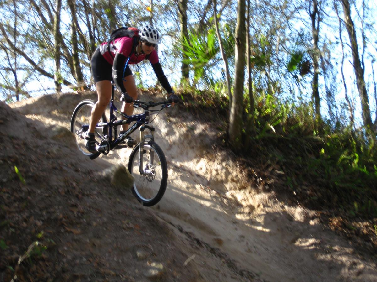 A mountain biker in a pink shirt is navigating a sandy slope on a forest trail, wearing a helmet and protective gear. Sunlight filters through the trees in the background, highlighting the natural surroundings. Alafia River State Park mountain bike trail.