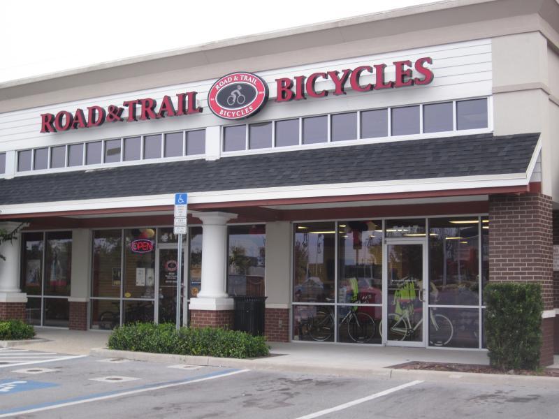 Exterior view of a bicycle shop named "Road & Trail Bicycles," featuring a red and white sign, large windows, and a clearly visible "OPEN" sign. The entrance showcases bicycles inside, with parking spaces in front and a landscaped area nearby.
