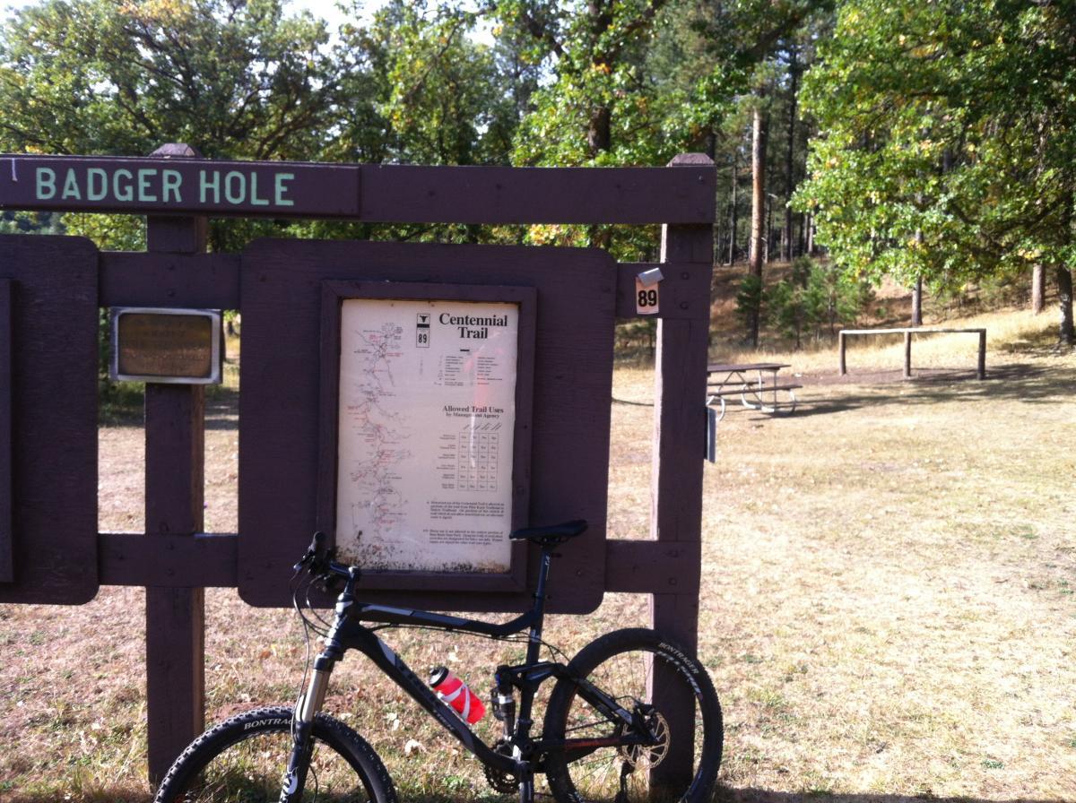 Signage at Badger Hole Trailhead, featuring a map of the Centennial Trail and allowed trail uses, with a mountain bike parked nearby in a grassy area surrounded by trees. Centennial Trail mountain bike trail.