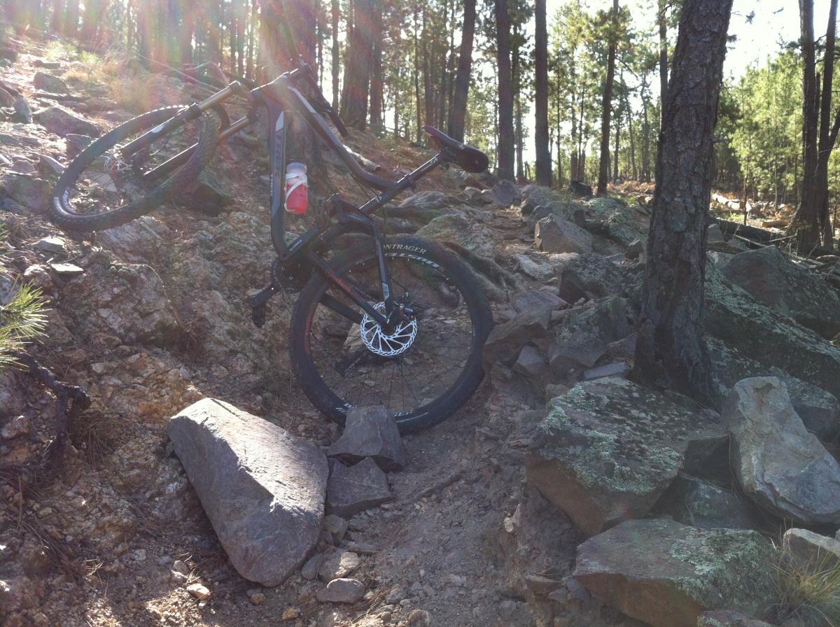 A mountain bike resting on a rocky trail in a forested area, with sunlight filtering through the trees. The bike has a water bottle attached and is positioned near several large rocks along the path. Centennial Trail mountain bike trail.