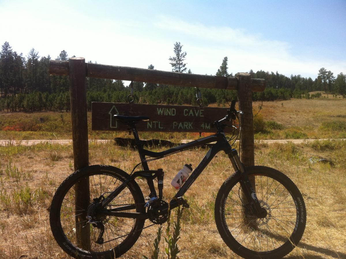 A mountain bike leaning against a wooden sign that points toward Wind Cave National Park, indicating a distance of 4 miles. The background features a grassy area with scattered trees under a clear blue sky. Centennial Trail mountain bike trail.