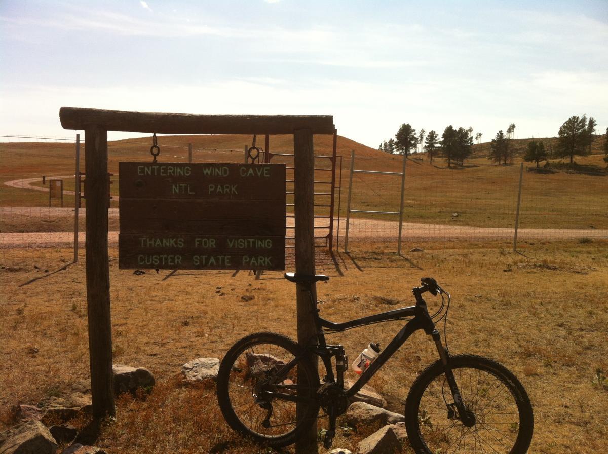 A mountain bike parked next to a wooden sign that reads "Entering Wind Cave Ntl Park" and "Thanks for visiting Custer State Park," set against a backdrop of open grassland and distant hills under a partly cloudy sky. Centennial Trail mountain bike trail.