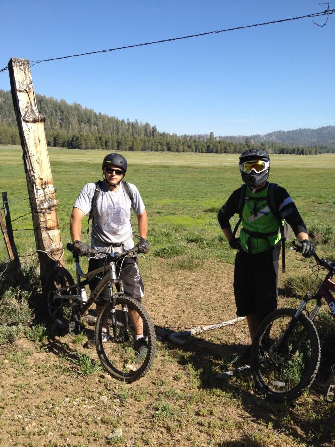 Two mountain bikers standing beside their bikes on a grassy field. One rider is wearing a gray t-shirt and helmet, while the other is dressed in a green and black outfit with a full-face helmet and goggles. A wooden fence and forested hills are visible in the background under a clear blue sky. The Cannell Plunge mountain bike trail.