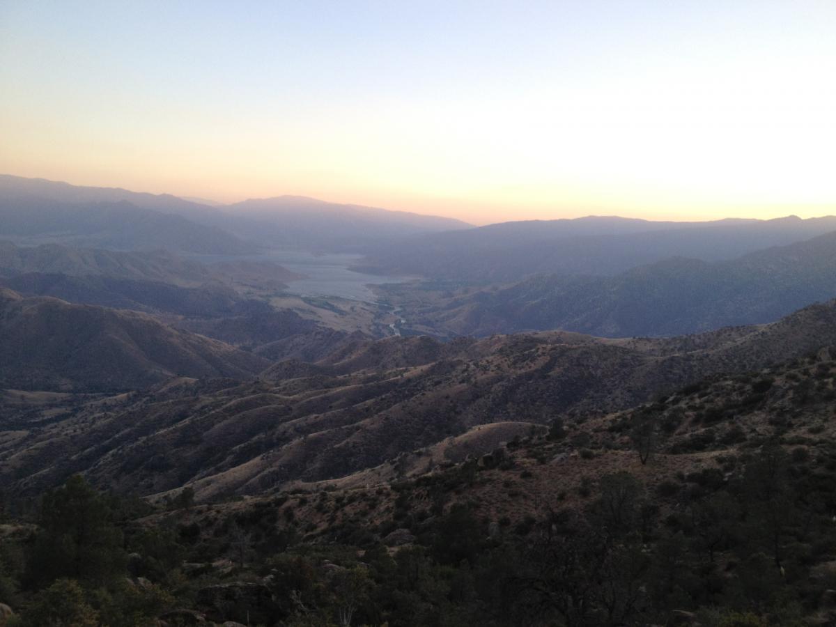 A panoramic view of rolling hills and mountains at dusk, with a soft gradient of colors in the sky transitioning from orange to blue. A river can be seen winding through the valley below, surrounded by greenery and rugged terrain. The Cannell Plunge mountain bike trail.