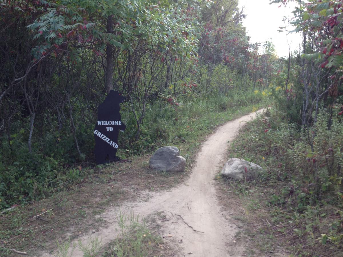 A narrow dirt path leads into a lush, green area with trees and bushes. On the left side of the path, a bear-shaped sign reads "WELCOME TO GRIZZLAND," welcoming visitors to the area. Large stones are scattered along the edge of the path, and the foliage is dotted with hints of autumn color. Elm Creek Park mountain bike trail.