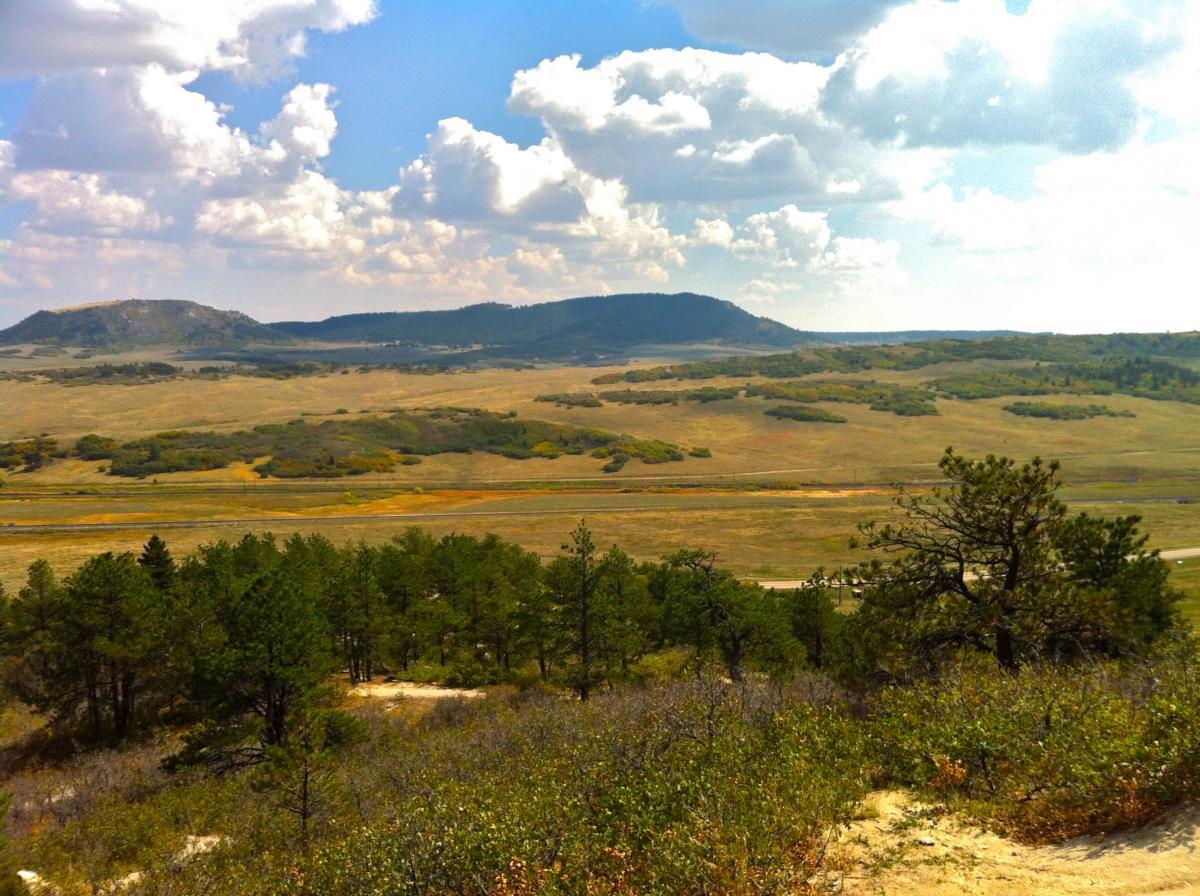 A panoramic view of a peaceful landscape featuring rolling hills, scattered patches of trees, and an expansive sky filled with clouds. The foreground includes greenery, while the background showcases distant mountains and open fields. Spruce Mountain Trail Upper Loop mountain bike trail.