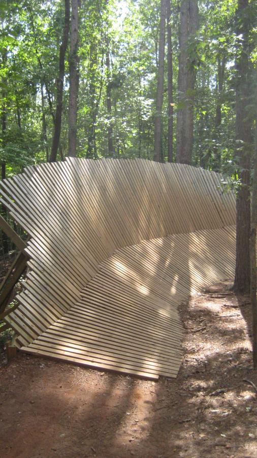 A winding wooden structure made of slats, set in a forested area. The path curves gently, leading through tall trees and dappled sunlight filtering through the leaves. The design appears to blend natural elements with a creative architectural feature. Issaqueena Lake mountain bike trail.