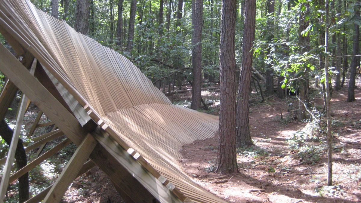 A wooden structure with slats arranged in a wave-like pattern, set within a forest of tall trees and greenery, partially shaded by the foliage above. The ground is covered with pine needles and small plants, creating a natural woodland atmosphere. Issaqueena Lake mountain bike trail.