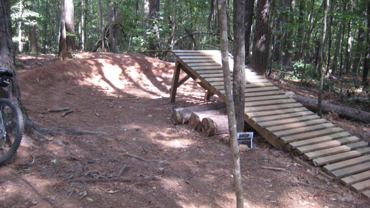 A wooden ramp leading up a small dirt jump in a forested area, surrounded by trees and pine needles. There is a bike partially visible on the left, and a sign indicating a drop-off area nearby. The setting is suitable for outdoor biking activities. Issaqueena Lake mountain bike trail.