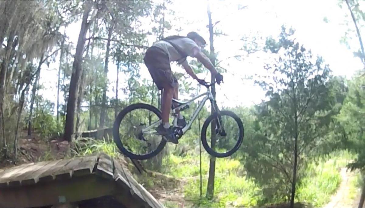 A young cyclist performing a jump off a wooden ramp in a forested area, with trees and greenery in the background. The rider is wearing a helmet and shorts, captured mid-air above the ramp. Santos mountain bike trail.
