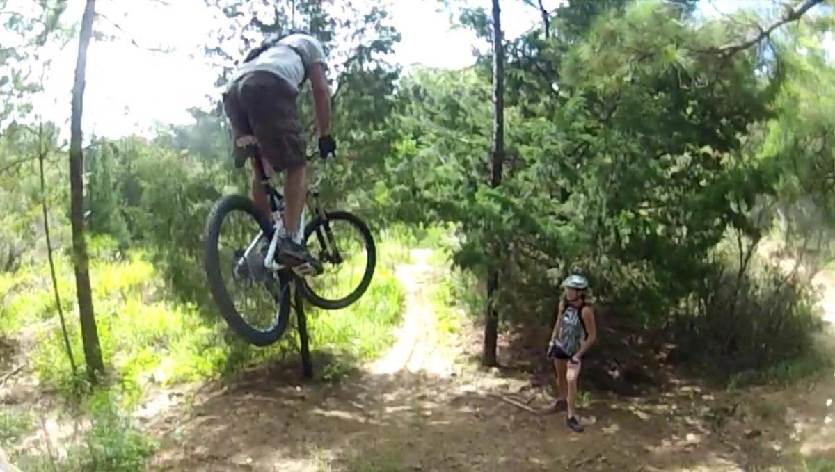 A mountain biker performs a jump on a dirt trail surrounded by trees, while a child watches nearby. The biker is wearing a gray shirt and shorts, and the child is wearing a helmet and casual clothing. The scene captures an outdoor biking adventure. Santos mountain bike trail.