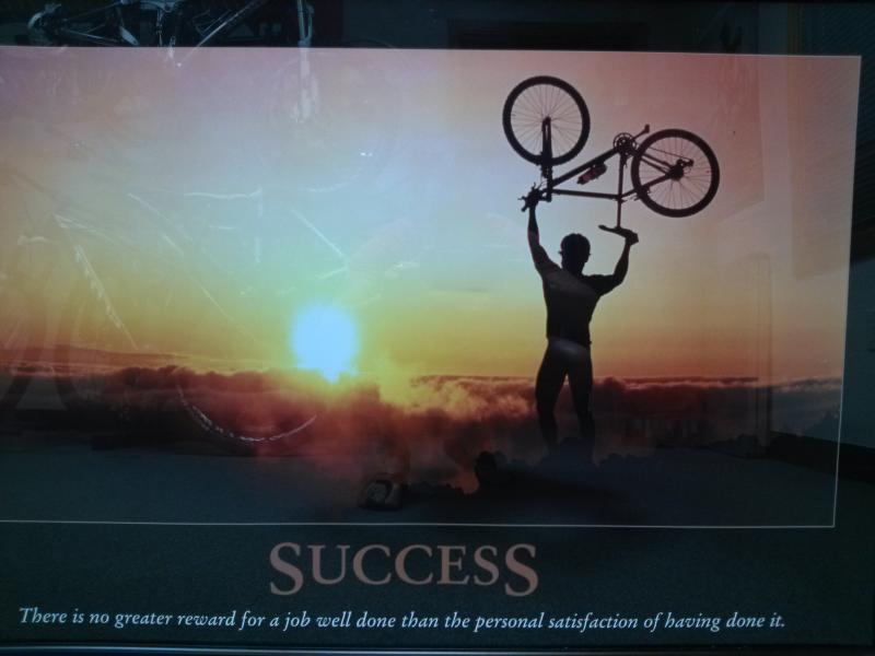 A silhouetted figure standing on an elevated surface raises a bicycle above their head against a vibrant sunset background. The word "SUCCESS" is prominently displayed, along with the quote: "There is no greater reward for a job well done than the personal satisfaction of having done it."